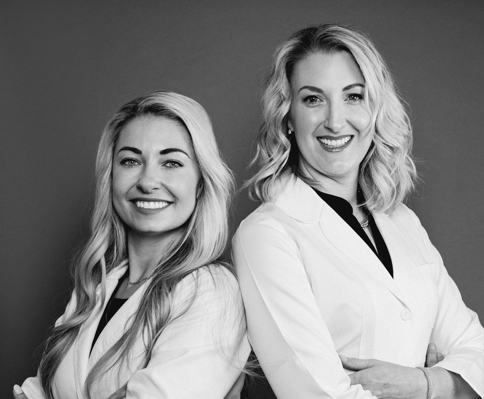 Black and white image of Dr. Lovell and Dr. Kile, of Urbana & Darby Dental Smiles, smiling and standing back to back in their dental coats.