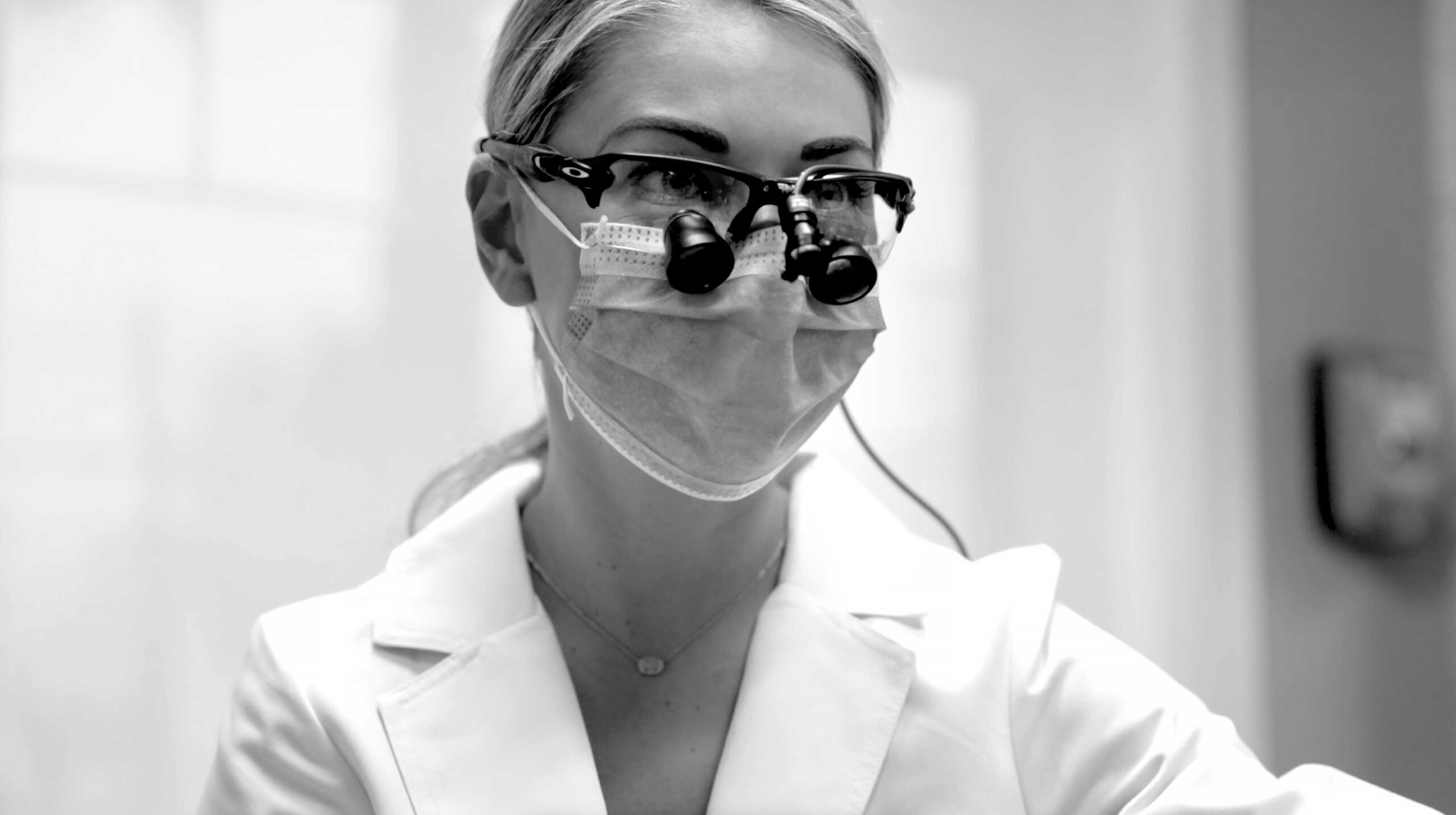 A black and white image of Dr. Lovell wearing a mask, dental glasses, and lab coat while in a dental exam room.