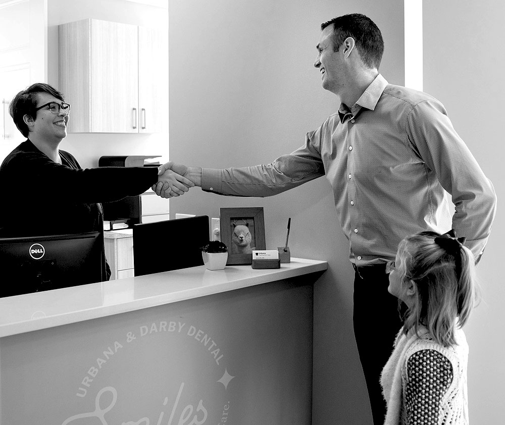 A black-and-white photograph of a man and his young daughter at the reception desk of Urbana & Darby Dental Smiles. The man is smiling and shaking hands with the receptionist, who is also smiling. The receptionist sits behind a desk, which has a small plant and a framed picture on it. The girl stands beside her father, looking up at the interaction.
