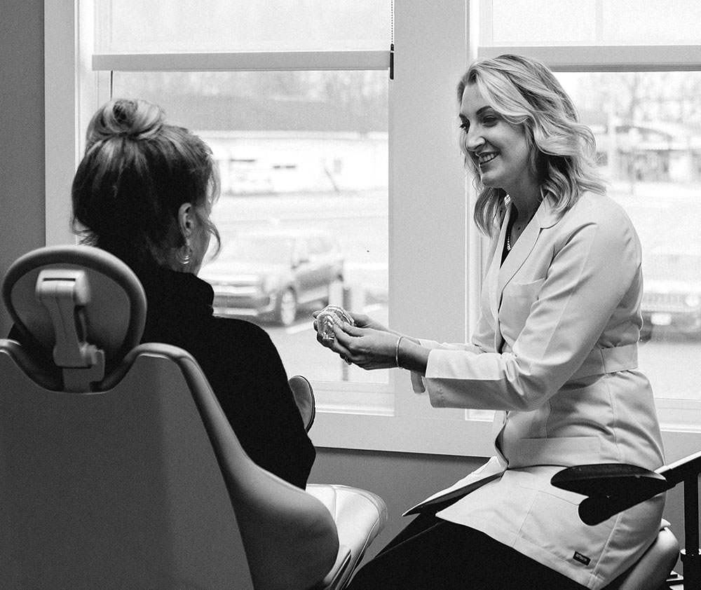 A black and white photo of Dr. Kile, from Urbana & Darby Dental Smiles, showing off a dental mouth model to a woman seated in a dental chair.